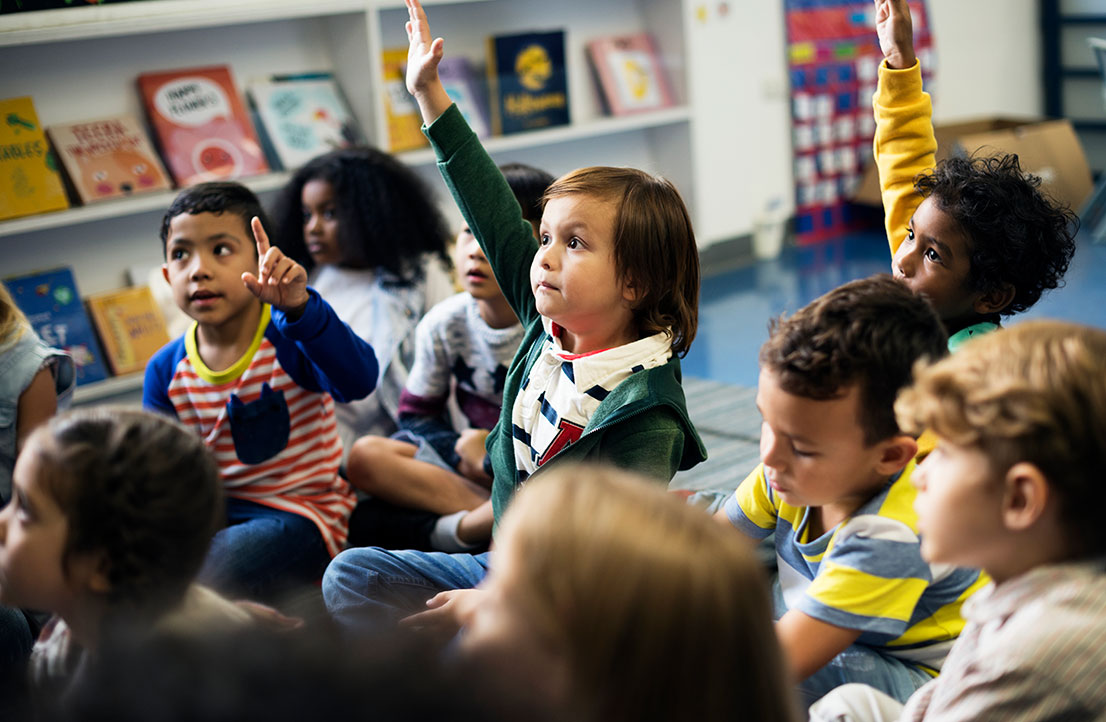 Children participating in an interactive learning session, raising their hands to engage in classroom activities as part of the Early Achievers program.